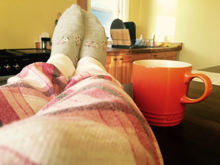 Feet on a kitchen table beside a Le Creuset mug in a sunny yellow kitchen