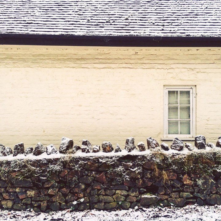 Stone cottage with dry stone wall in front of it covered in snow.