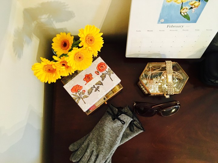 Top of a dressing table featuring calendar, flowers, jewellery box, gloves, sunglasses and perfume.
