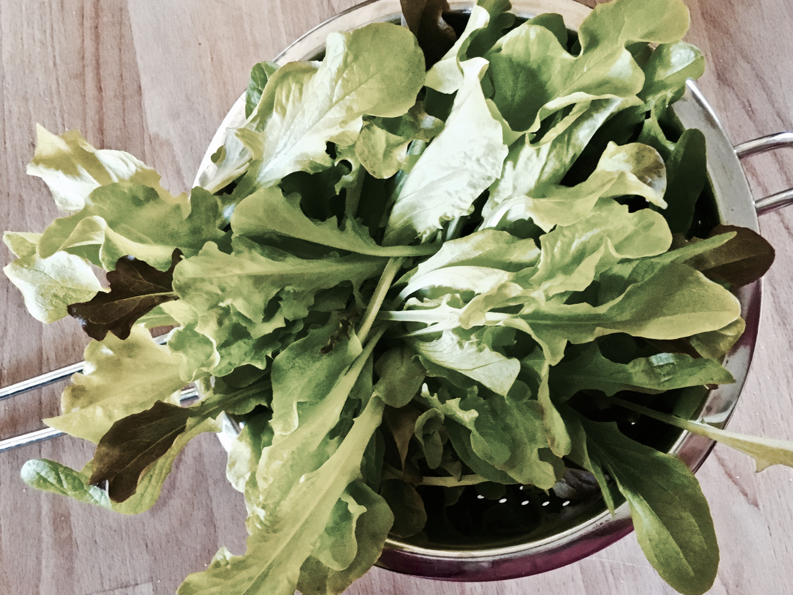 Colander of baby salad leaves on wooden chopping board.