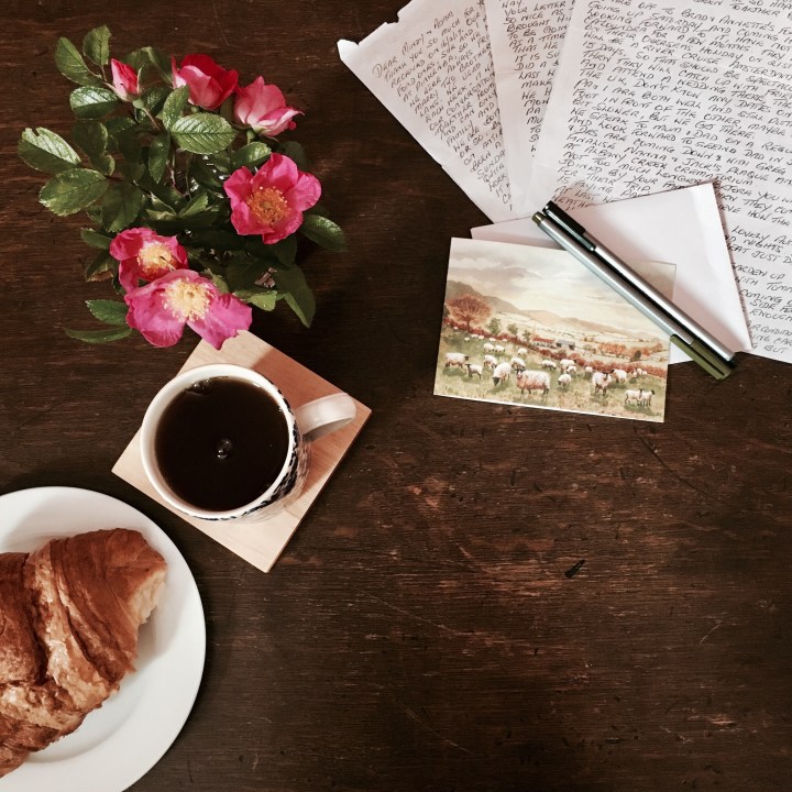 Flat lay of tea, croissant, roses, card and handwritten letter on timber table.