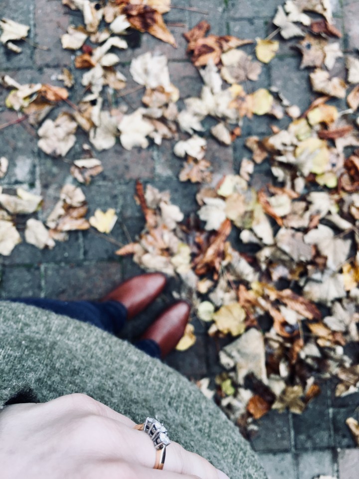 Engagement ring on woman wearing green jumper, jeans and boots surrounded by autumnal leaves