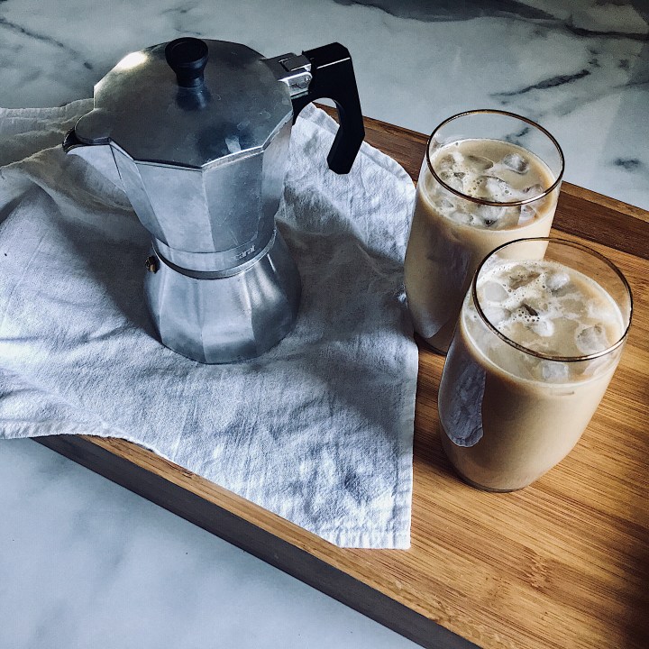 Tray on marble bench, where linen tea towel, stove top cafetiere and two glasses of iced coffee sit.