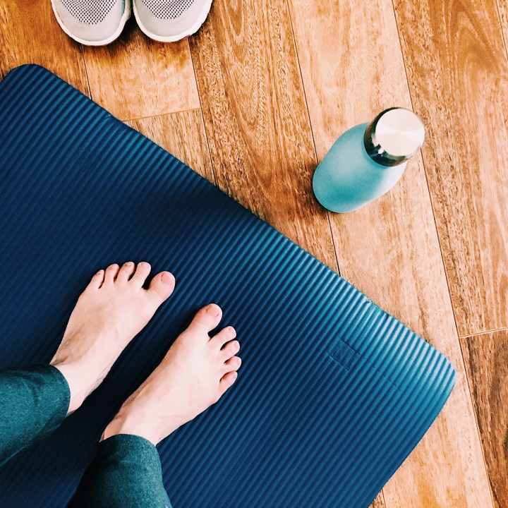 Woman wearing grey leggings standing on a blue yoga mat. A pair of sneakers and a blue water bottle are also in shot.