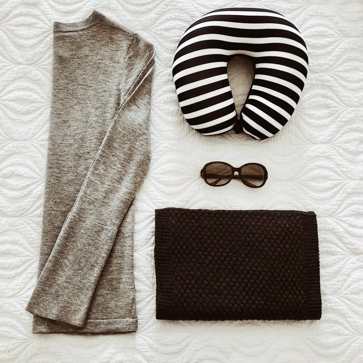 Flat lay of grey jumper, travel pillow, sunglasses and scarf.
