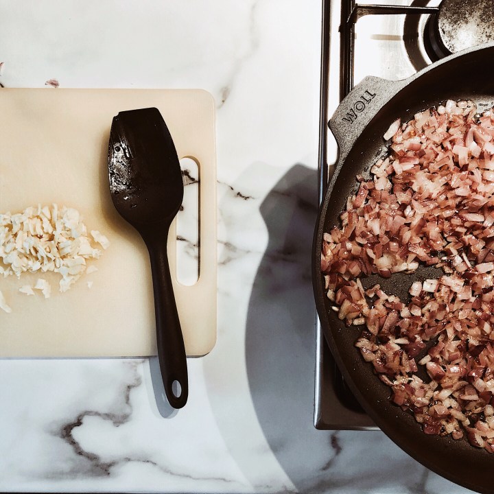 Red onions frying in a pan, with diced garlic on a chopping board besides.