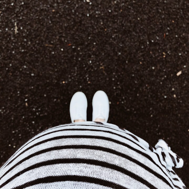 Pregnant woman in black and white stripe shirt looking at white sneaker clad feet.