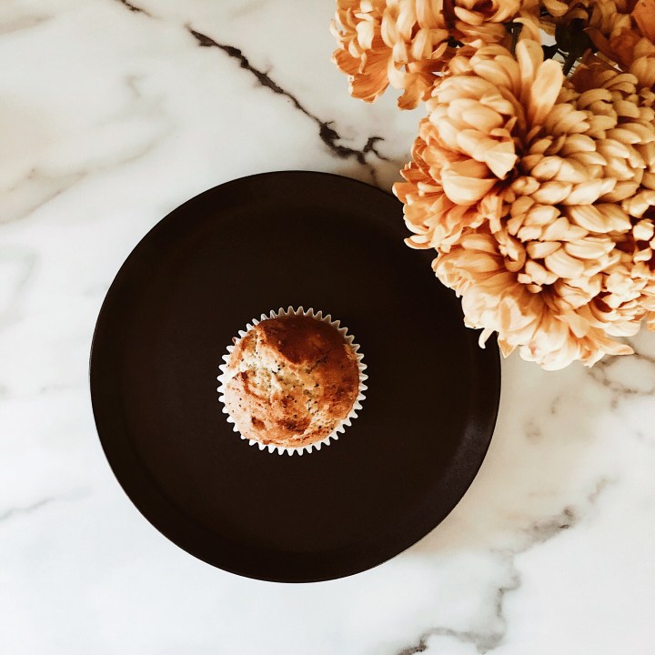 Orange and poppyseed muffin in the centre of a black plate beside a jar of yellow dahlias.