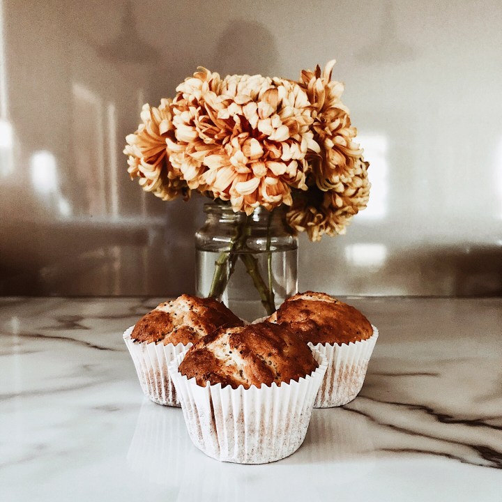 Yellow dahlias in a jar beside three orange and poppyseed muffins.