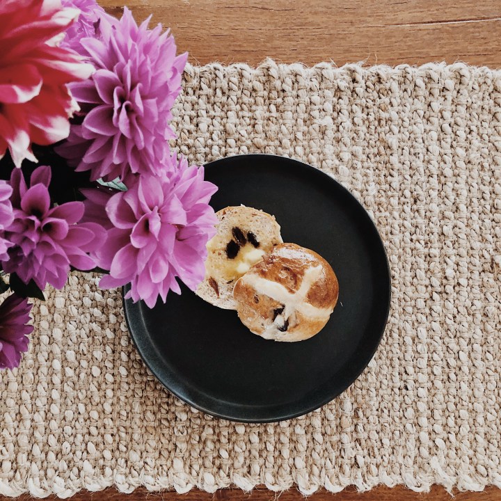 Warm hot cross bun with butter underneath a vase of red and purple dahlias.