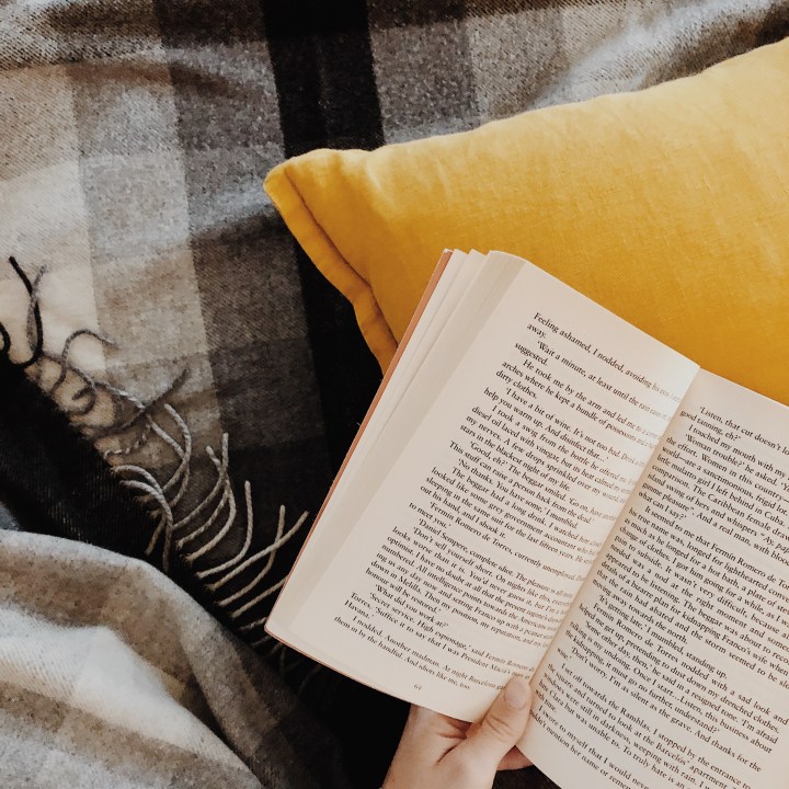 Hand holding a book surrounded by a black and white check wool blanket and mustard yellow linen cushion.