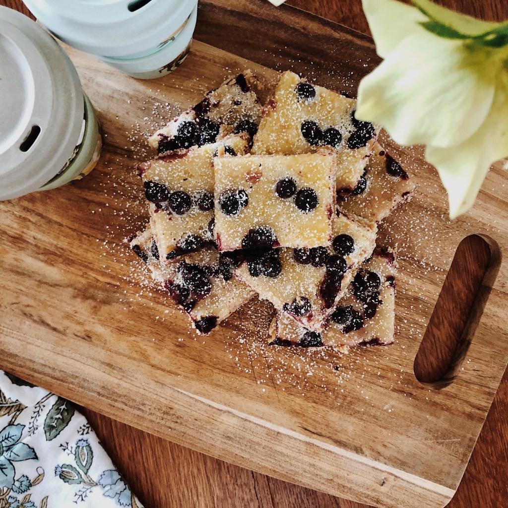 Lemon and blueberry squares dusted with icing sugar sitting on a wooden board, besides two reusable coffee cups and underneath a vase of ellebores.