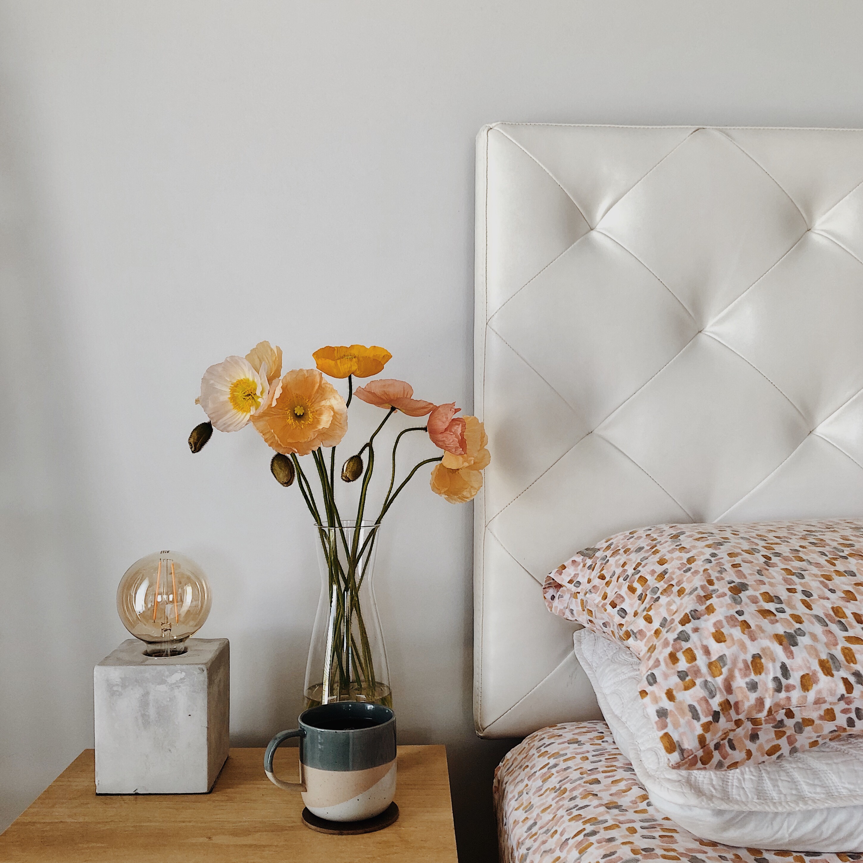 Bedside table wit lamp, mug and vase of poppies besides a made bed.