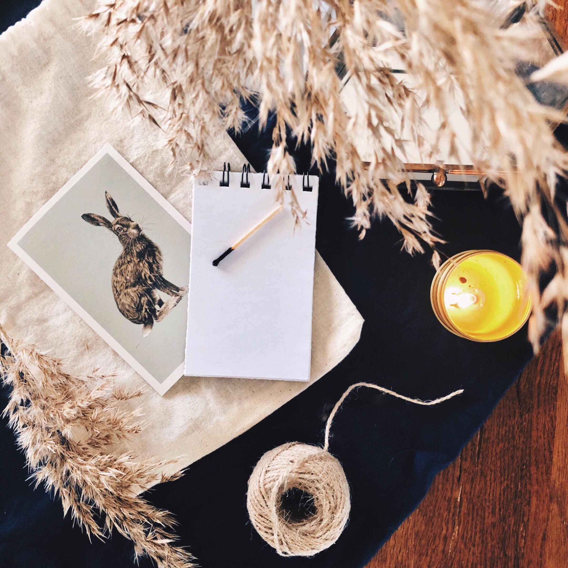 Linen, cotton, dried grasses, a notebook, candle, string and vintage postcard sitting on a timber table.