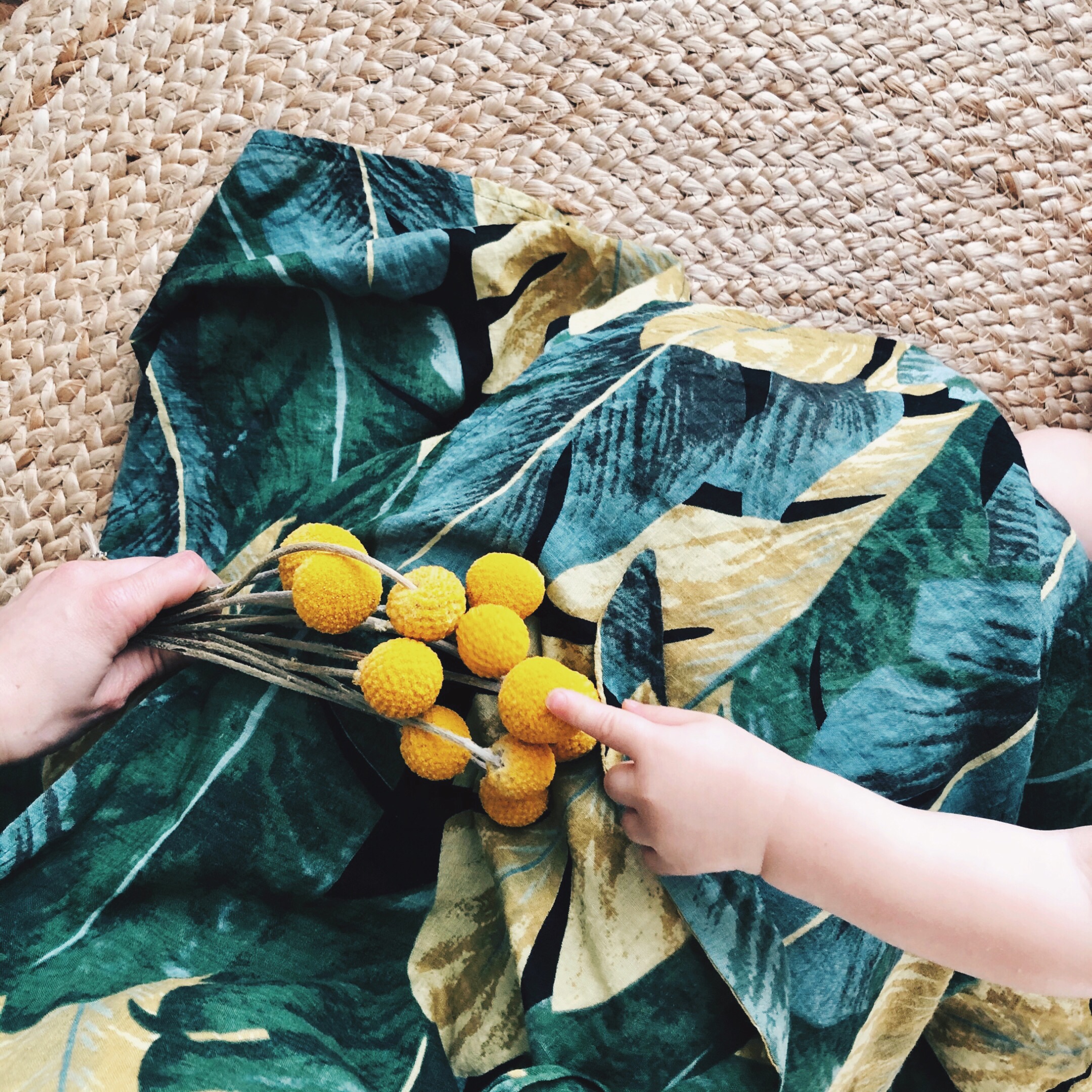 Woman sitting on jute rug holding bunch of billy button flowers, which are being poked by a small toddler hand.