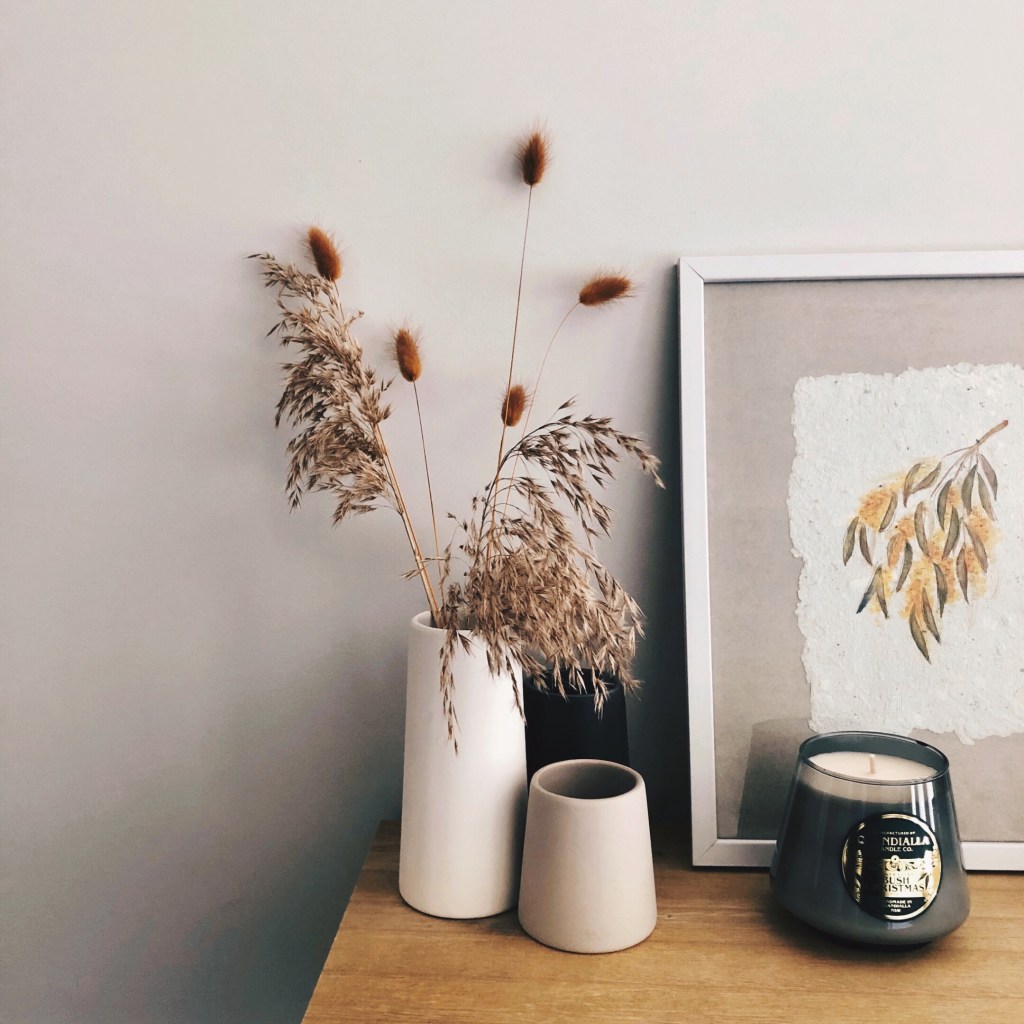 Candle sitting on a table beside vases of dried grasses and a hand painted drawing of wattle.