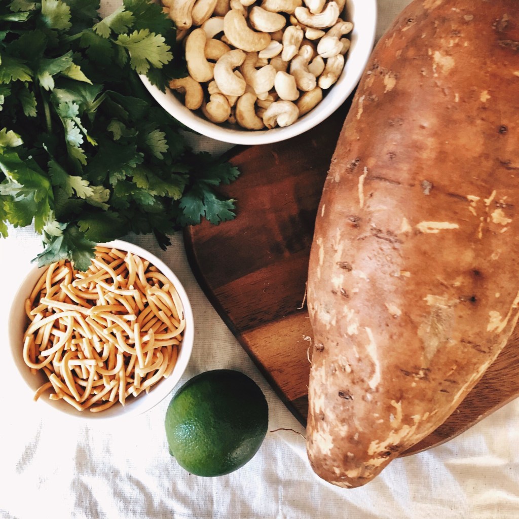 Sweet potato on a wooden board, surrounded by fried noodles, cashews, coriander and a lime.