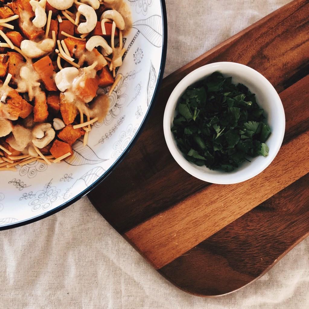 Sweet potato salad in a large bowl, beside a small bowl of chopped coriander, sitting on a wooden chopping board.