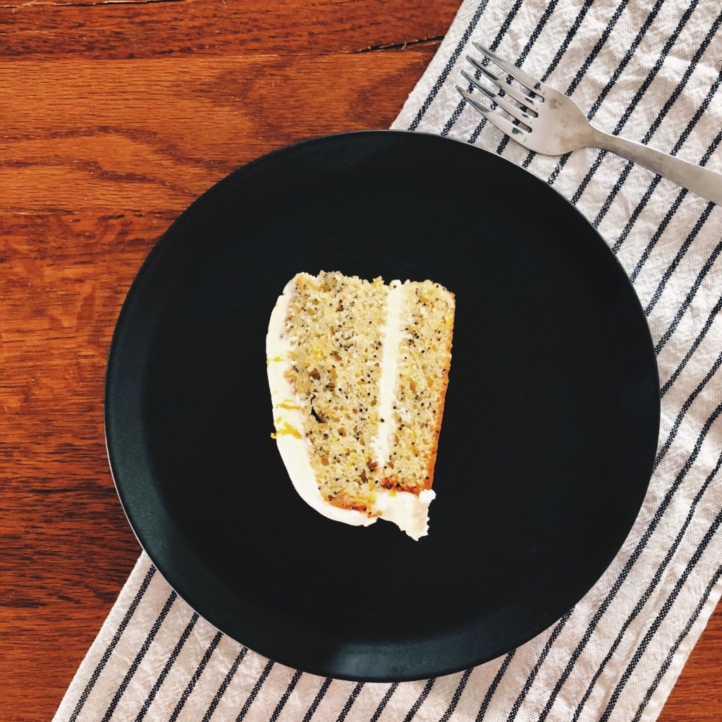 A slice of orange and poppyseed cake on a black plate, atop a wooden table and black and white stripe tea towel.