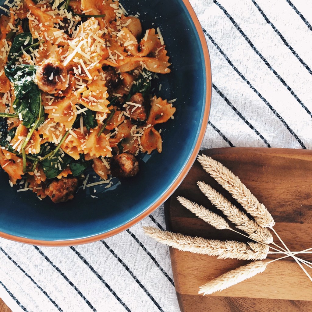 Pesto pasta with meatballs in a bowl, sitting on a striped tea towel beside a board with some ornamental dried grasses..