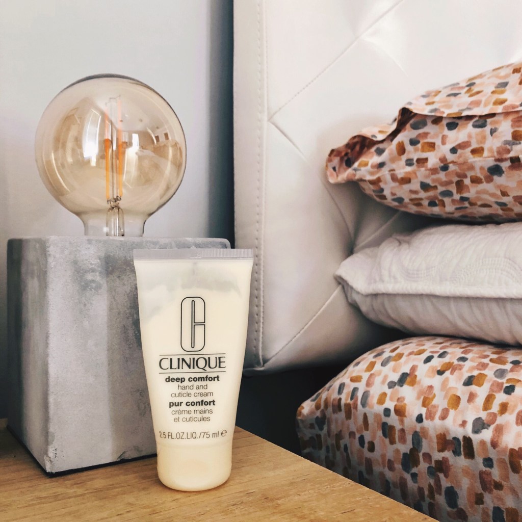 Clinique Deep Comfort Hand and Cuticle Cream in front of a lamp on a table beside a made bed.