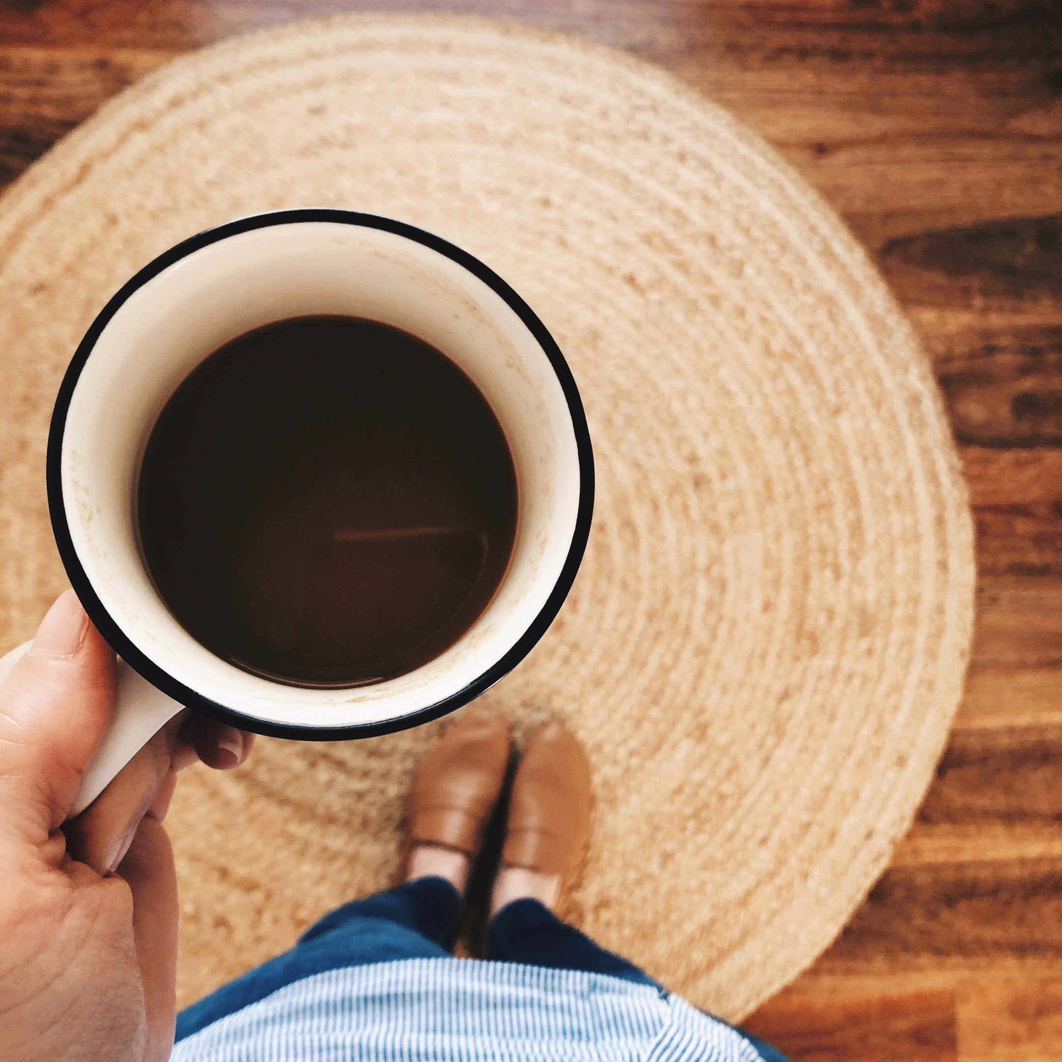 Woman in tan leather loafers, jeans and blue and white striped shirt standing on a jute rug holding a cup of coffee.
