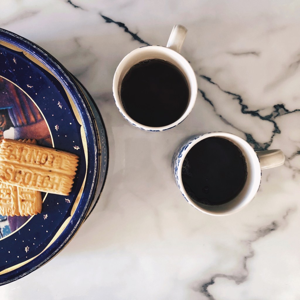 Scotch fingers atop a biscuit tin beside two cups of tea.