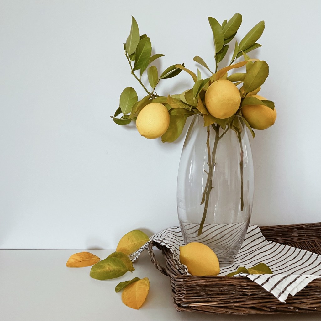 Lemon laden branches in a tall glass vase on a wicker serving tray lined with a black and white striped tea towel.