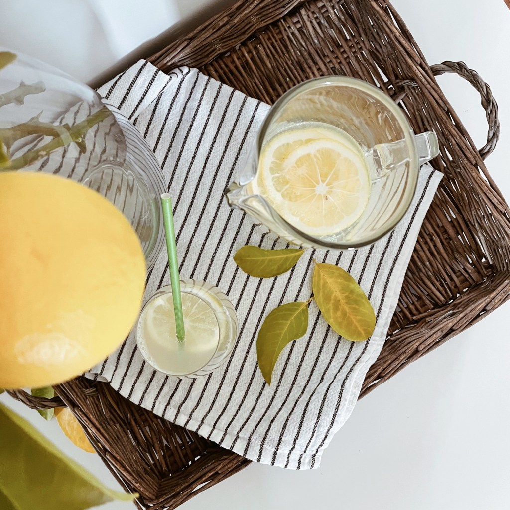Home made lemonade in a jug and glass, sat on a wicker serving tray with a striped tea towel, beside a jar of lemon laden branches.