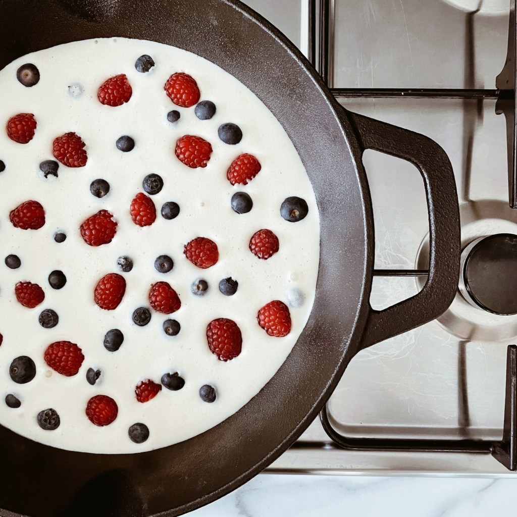 Looking down at clafoutis mixture in a cast iron skillet, dotted with blueberries and raspberries.