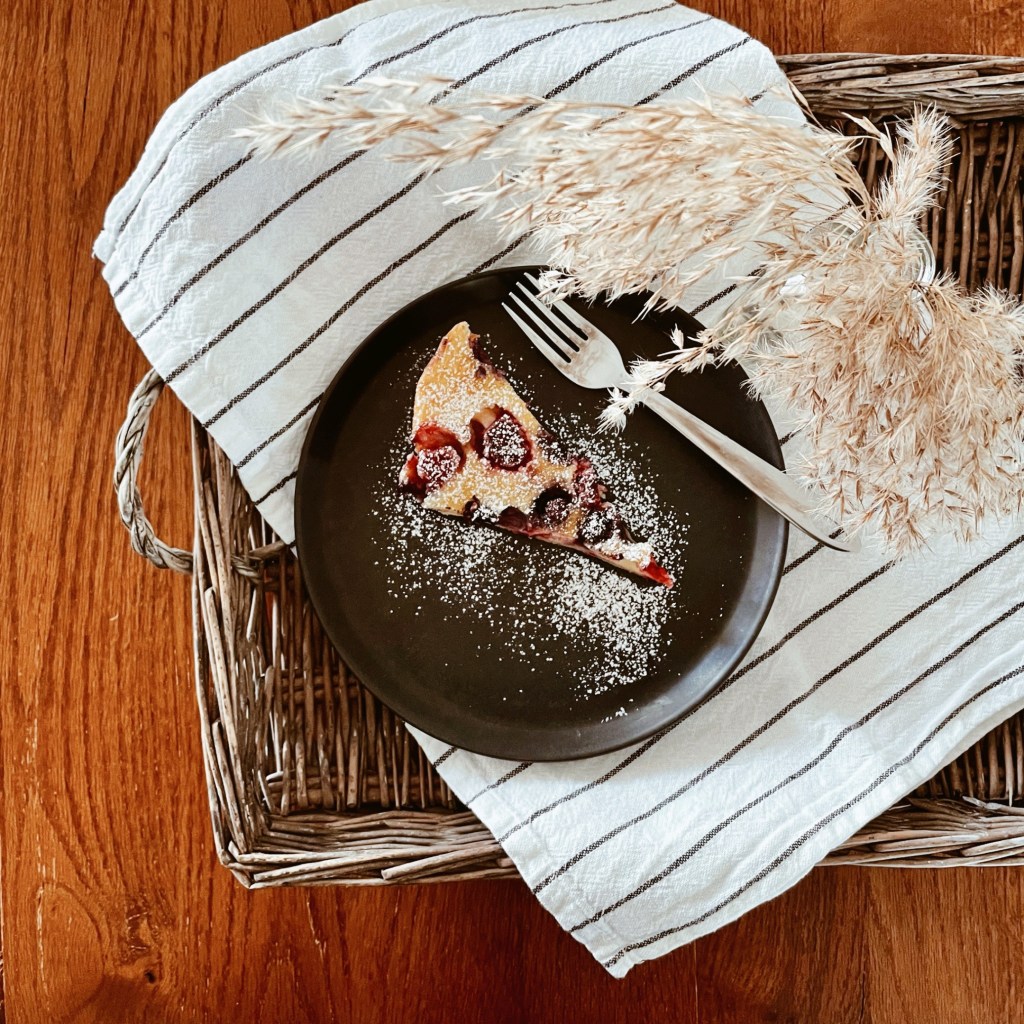 Berry clafoutis on a black plate sitting on a black and white striped teatowel in a wicker tray. A vase of dried grasses is beside.