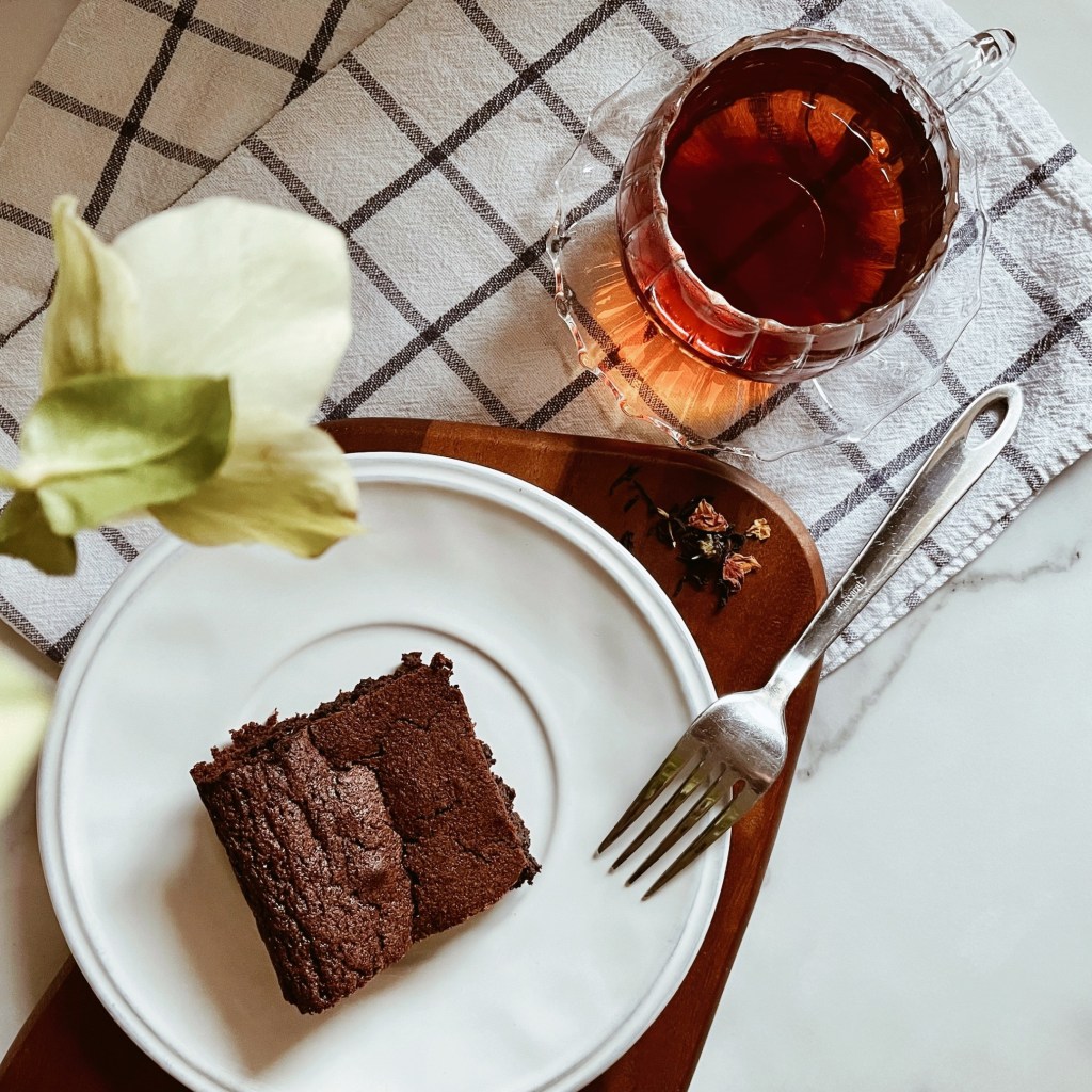 Looking down thrown arranged hellebores to a chocolate brownie and cup of tea arranged on a wooden board and checked teatowel.