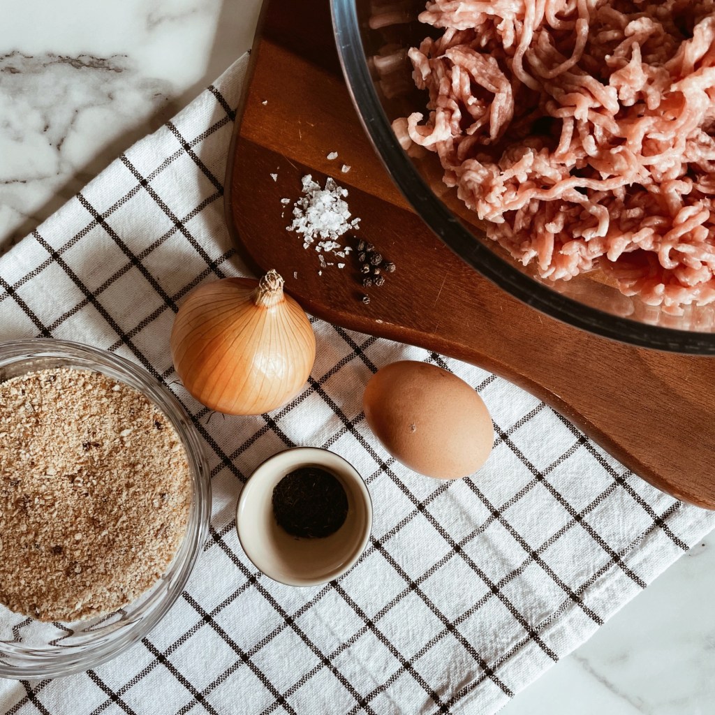 Pork mince in bowl, besides an onion, egg and breadcrumbs and dill in two separate smaller bowls. 