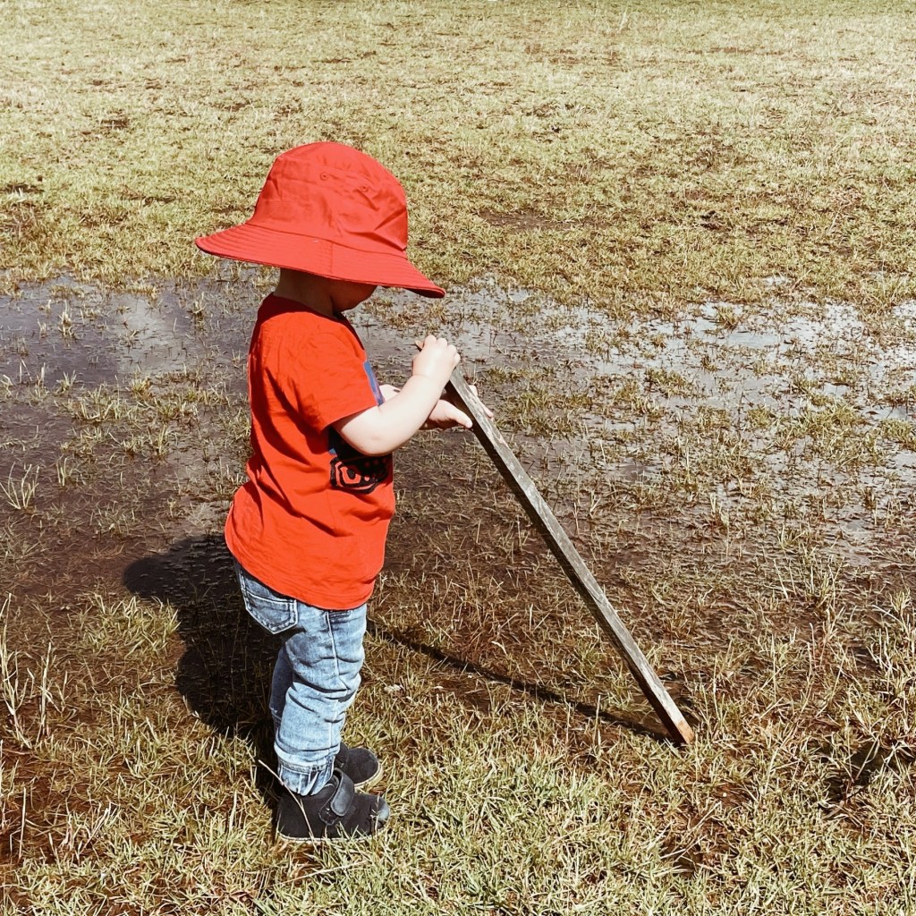 Toddler in oversized hat standing by a puddle and holding a stick.