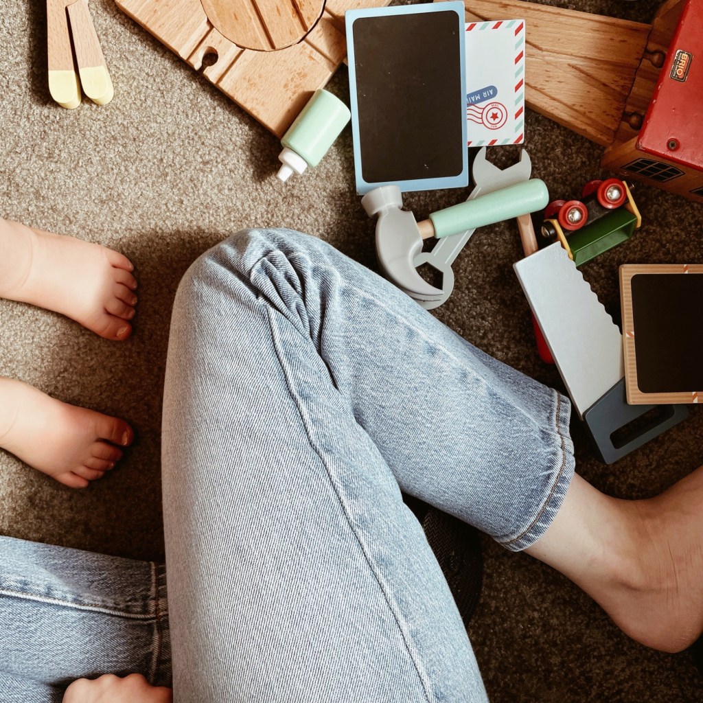 Looking down at a woman's legs, toddler feet and wooden toys scattered everywhere.