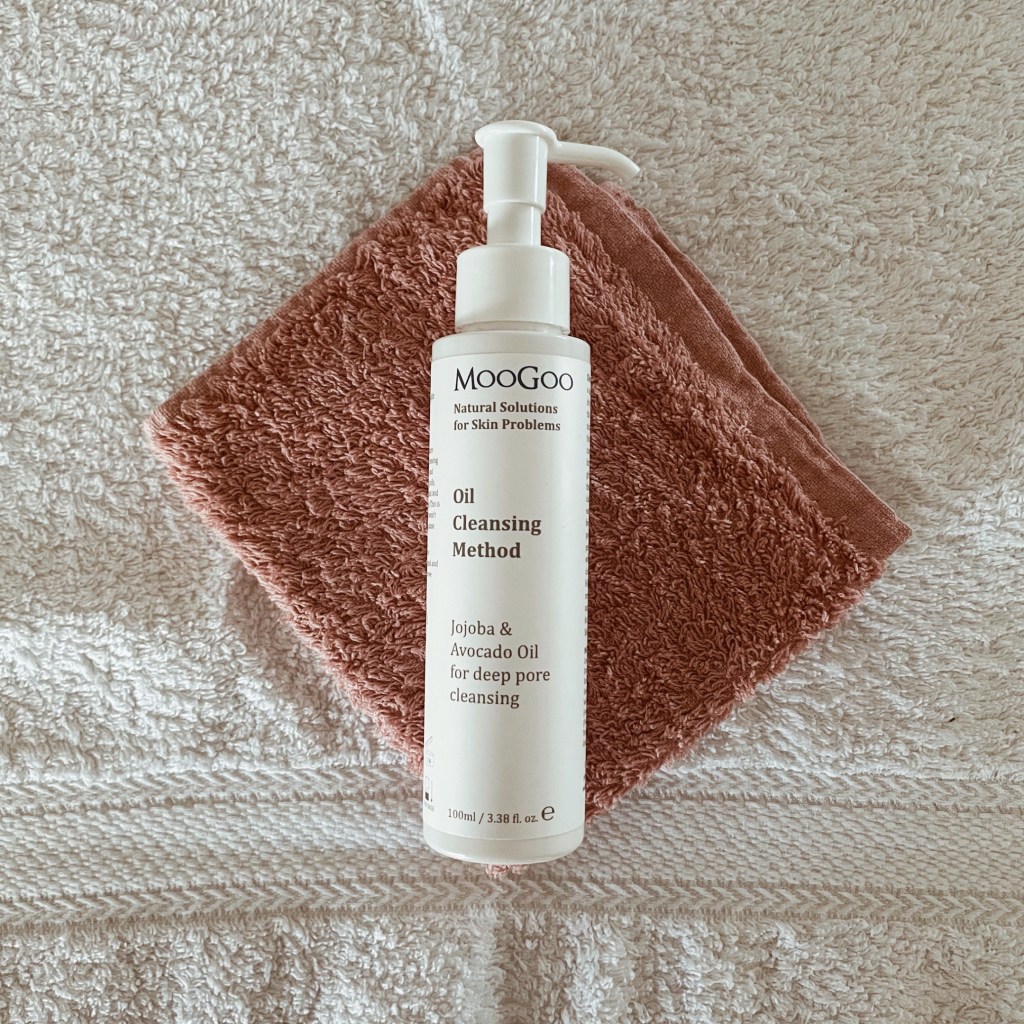 MooGoo Oil Cleansing Method bottle on pink face washer against white towel.