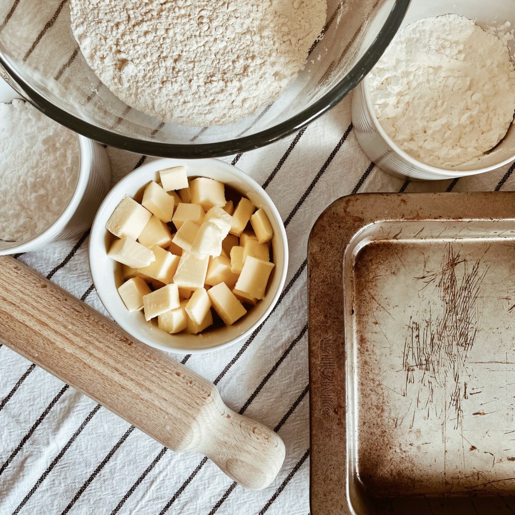 Bowls and ramekins of flour, cornflour, icing sugar and butter beside an old baking tin and wooden rolling pin.
