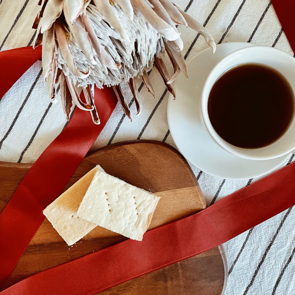 Looking down on two slices of shortbread on a wooden board, surrounded by a red ribbon, dried protea and cup of tea.