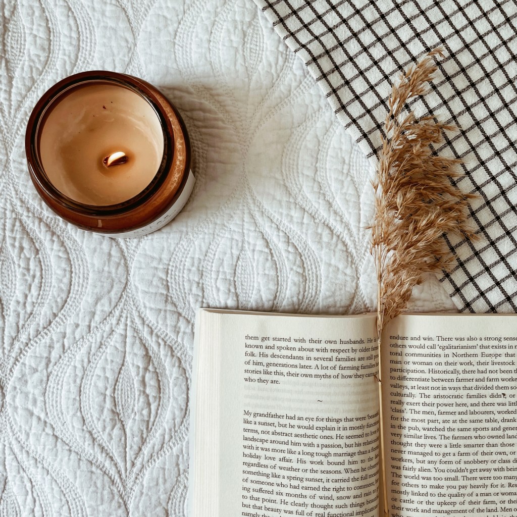 Looking down on a candle, book and blade of feathered grass atop a white coverlet and black and white check tea towel.